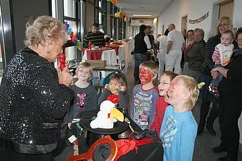 Eine ältere Frau mit glitzerndem Oberteil unterhält eine Gruppe von Kindern, die mit Gesichtern bemalt sind. Die Kinder schauen gespannt zu, während die Frau einen Zauberhut und Requisiten präsentiert. Im Hintergrund sind weitere Personen und festliche Dekorationen zu sehen.