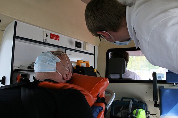 Ein Patient liegt in einem Krankenwagen auf einer Trage. Der Patient trägt eine Gesichtsmaske und hat seinen Kopf zurückgelehnt. Ein Sanitäter beugt sich über ihn, während er mit ihm spricht. Im Hintergrund sieht man weitere medizinische Ausstattungen.