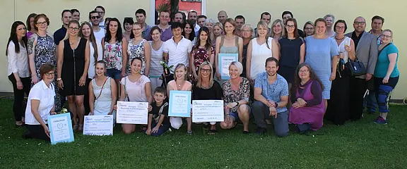 Große Gruppe lächelnder Erwachsener posiert im Freien vor einer hellen Wand. In der ersten Reihe halten mehrere Personen Urkunden und große Schecks. Die Szene wirkt wie eine Preisverleihung oder Abschlussfeier im Pflege- oder Bildungsbereich.