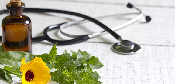 A stethoscope rests beside a small brown medicine bottle and fresh green herbs with a yellow flower on a white wooden surface, suggesting natural or herbal remedies alongside conventional healthcare.