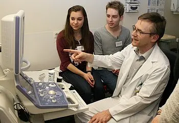 Ein Arzt im weißen Kittel erklärt etwas an einem Ultraschallgerät. Zwei Jugendliche, ein Mädchen und ein Junge, hören aufmerksam zu und schauen auf den Bildschirm. Der Arzt zeigt mit dem Finger auf eine Stelle auf dem Bildschirm.