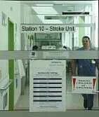 Krankenhausflur mit Glastüren, beschriftet „Station 10 – Stroke Unit“. Dahinter medizinisches Personal auf dem Gang; Hinweis- und Besucherschilder am Eingang.