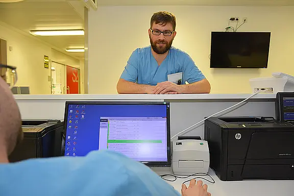 Krankenhauspersonal an der Anmeldung: Eine Pflegekraft sitzt am Computer, eine weitere steht am Tresen und spricht mit ihm. Auf dem Monitor ist eine Liste geöffnet. Szene zeigt Arbeitsabstimmung am Stationsstützpunkt.