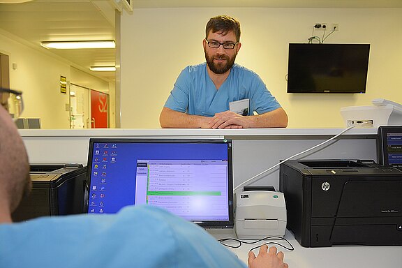 Krankenhauspersonal an der Anmeldung: Eine Pflegekraft sitzt am Computer, eine weitere steht am Tresen und spricht mit ihm. Auf dem Monitor ist eine Liste geöffnet. Szene zeigt Arbeitsabstimmung am Stationsstützpunkt.