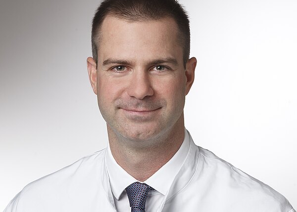 Nahaufnahme eines Mannes mit kurzem Haar, leichtem Lächeln, in weißem Hemd und dunkel gemusterter Krawatte vor neutralem Hintergrund.