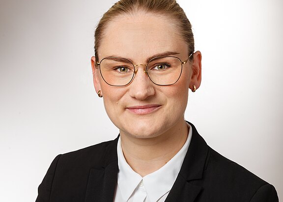 Porträtfoto einer Person in schwarzem Blazer und weißem Hemd, mit runder Brille, nach vorn blickend und leicht lächelnd, vor neutralem, hellem Hintergrund.