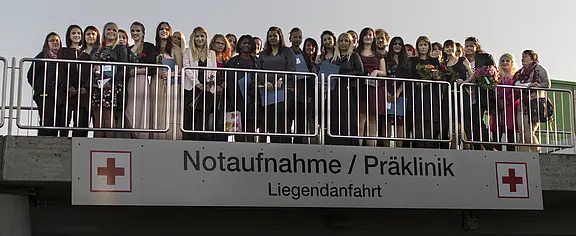 Große Gruppe von Menschen posiert hinter einem Geländer über einem Schild „Notaufnahme/Präklinik – Liegendanfahrt“ mit roten Kreuzsymbolen; Szene vor einer Krankenhauszufahrt.