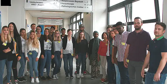Gruppe junger Erwachsener steht in einem Krankenhausflur. Sie tragen Besucher- oder Namensschilder und blicken zur Kamera. Über ihnen hängen Wegweiser zu Abteilungen. Eindruck: gemeinsamer Rundgang oder Einführung.