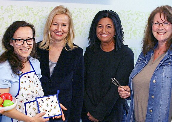 Vier Frauen stehen lächelnd nebeneinander. Links trägt eine Frau eine Schürze, hält eine Schüssel mit buntem Gemüse und zwei Topflappen. Daneben halten zwei Frauen Küchenutensilien, eine davon einen Schneebesen. Die Szene wirkt wie ein gemeinsames Koch- oder Küchenprojekt.