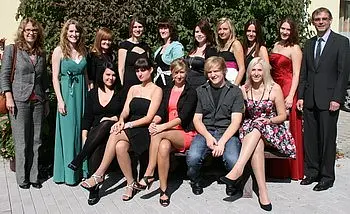 Die Gruppe besteht aus 15 Personen, die hellere und dunklere Kleidung tragen, darunter elegante Kleider und Anzüge. Sie posieren gemeinsam im Freien, umgeben von Pflanzen. Die Stimmung scheint festlich und freundschaftlich zu sein.