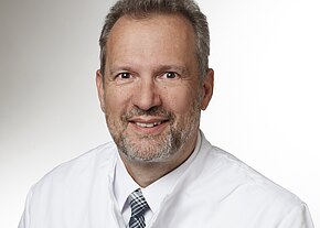 Univ.-Prof. Dr. med. Hans Jürgen Heppner, MHBA Porträt eines lächelnden Mannes mittleren Alters im weißen Arztkittel mit kariierter Krawatte, vor neutralem Hintergrund. Nahaufnahme, Kopf und Schultern, direkter Blick in die Kamera.