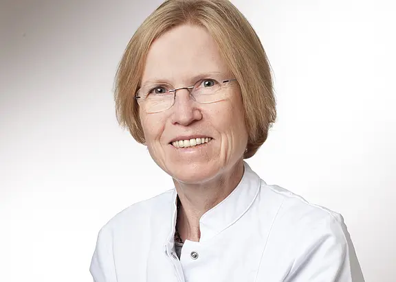 Porträt einer freundlich lächelnden Person mit Brille und kurzem, hellblondem Haar, in weißem Arztkittel vor hellem, neutralem Hintergrund.
