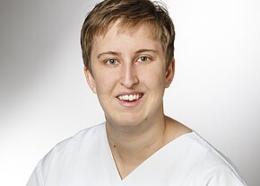 Station 3b Porträt einer lächelnden Person mit kurzem Haar in weißem Kasack, frontal aufgenommen vor hellem, neutralem Hintergrund. Der Fokus liegt auf dem freundlichen Gesicht.