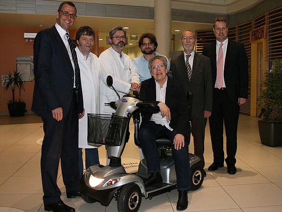 Sieben Erwachsene posieren in einem hellen Foyer. Ein Mann sitzt auf einem silbernen Elektromobil, die anderen stehen daneben, einige in weißen Kitteln, andere in Anzügen. Die Gruppe wirkt wie Ärzte und Verantwortliche, die das Mobilitätshilfsmittel präsentieren.