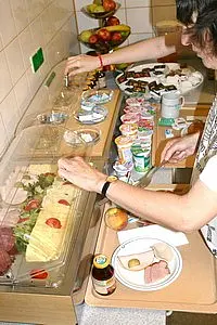 Eine Person steht an einem Buffet und wählt verschiedene Lebensmittel aus. Auf dem Tisch befinden sich Aufschnitt, Käse, frisches Obst und Joghurt. Die Person füllt einen Teller mit Lebensmitteln und greift nach einem Getränk. Im Hintergrund sind weitere Speisen und eine Schale mit Äpfeln zu sehen.
