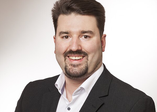 Porträtfoto eines lächelnden Mannes mit dunklem Haar und kurzem Bart, im dunklen Sakko und weißen Hemd, vor hellem neutralem Hintergrund. Kopf und Oberkörper frontal, professionelle Aufnahme.