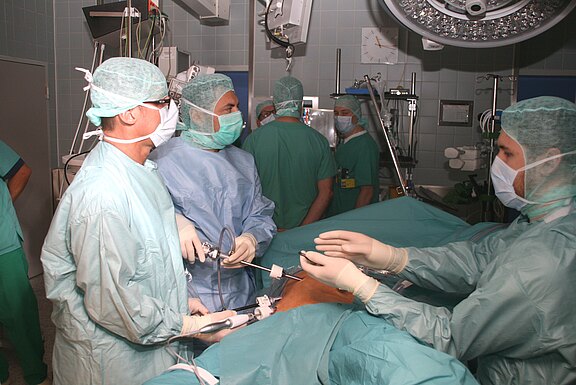 Operationsteam im OP-Saal führt eine minimalinvasive Bauchoperation durch. Mehrere Chirurgen in steriler Kleidung bedienen Laparoskop und Instrumente an einem Patienten unter OP-Licht; Monitore und Geräte im Hintergrund.