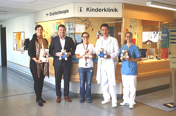 Fünf Erwachsene stehen vor dem Empfang der Kinderklinik im Krankenhaus und halten Teddybären in den Händen, als Zeichen einer Spielzeugspende oder Übergabe an die Kinderabteilung.
