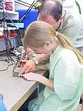 Ein Erwachsener zeigt einem Kind, wie man an einem technischen Gerät arbeitet. Beide konzentrieren sich auf die Arbeit an einem Tisch, während verschiedene Werkzeuge und Kabel im Hintergrund sichtbar sind. Das Kind trägt einen grünen Kittel und scheint aufmerksam zu lernen.