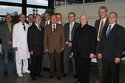 Eine Gruppe von zwölf Männern steht in einem modernen Raum. Einige tragen Anzüge, einer ist in einem weißen Kittel. Die Männer lächeln und wirken freundlich, während sie sich in zwei Reihen aufstellen. Pflanzen und große Fenster sind im Hintergrund sichtbar.