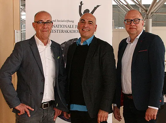 Drei Männer in Business-Kleidung stehen nebeneinander und lächeln in die Kamera. Hinter ihnen ein Informationsbanner; die Szene wirkt wie ein offizielles Treffen oder eine Veranstaltung in einem modernen Innenraum.