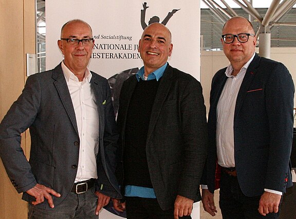 Drei Männer in Business-Kleidung stehen nebeneinander und lächeln in die Kamera. Hinter ihnen ein Informationsbanner; die Szene wirkt wie ein offizielles Treffen oder eine Veranstaltung in einem modernen Innenraum.