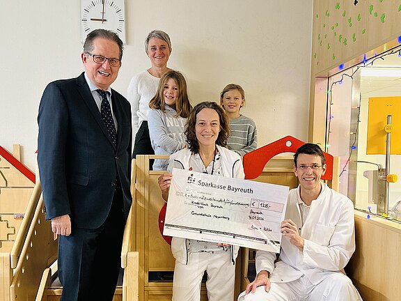 In einem hellen Kinderbereich einer Klinik posieren Mitarbeitende in weißer Kleidung mit einem großen Spendenscheck. Weitere Erwachsene und zwei Kinder stehen daneben und lächeln.