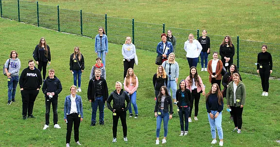 Eine Gruppe von 30 Personen steht auf einer grünen Wiese. Sie sind lächelnd angeordnet, einige tragen Jacken und Hoodies. Im Hintergrund ist ein Zaun sichtbar. Es ist eine freundliche, gesellige Stimmung.