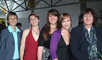 Fünf Frauen stehen nebeneinander in formeller Kleidung. Sie lächeln und scheinen bei einer Veranstaltung zusammen zu sein. Jede hat einen eigenen Stil, von elegant bis lässig, und die Atmosphäre wirkt freundlich und feierlich.