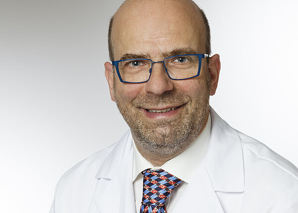 Porträt eines lächelnden Arztes in einem weißen Kittel, mit blauen Brillen und einem bunten, gemusterten Schlips. Der Hintergrund ist hell und neutral, wodurch der Fokus auf dem Arzt liegt.