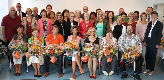 Gruppenfoto in einem Raum: Rund dreißig Erwachsene posieren. In der ersten Reihe sitzen mehrere Geehrte und halten große Blumensträuße, umgeben von lächelnden Kolleginnen und Kollegen. Anlass wirkt wie eine Ehrung oder Verabschiedung.