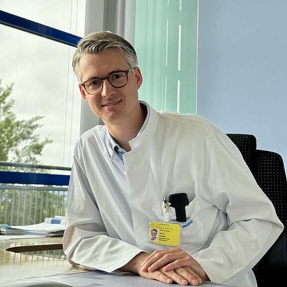 Ein Arzt in einem weißen Kittel sitzt an einem Schreibtisch. Er trägt eine Brille und lächelt freundlich in die Kamera. Auf seinem Kittel ist ein Namensschild sichtbar. Im Hintergrund sind Fenster und ein unscharfer Blick ins Freie.