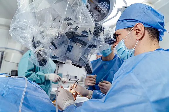 Chirurgenteam im OP führt eine präzise Mikrochirurgie durch. Der leitende Chirurg blickt durch ein steril abgedecktes Operationsmikroskop, während Hände Instrumente am Patienten führen. Schwerpunkt: moderne, hochpräzise Operation mit Teamarbeit und steriler Umgebung.