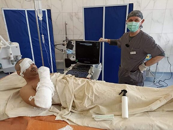 Ein Patient mit Verbänden auf dem Kopf und Arm liegt auf einem Bett in einem Krankenhauszimmer. Neben ihm steht ein lächelnder Arzt in Arbeitskleidung und Mundschutz, der an einem Ultraschallgerät arbeitet. Die Atmosphäre wirkt medizinisch und professionell.