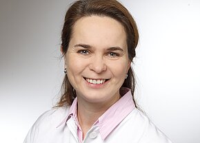 Dr. med. Claudia Anhut Porträt einer lächelnden Frau mit langen, braunen Haaren, die ein weißes Hemd mit rosa Kragen trägt. Der Hintergrund ist hell und neutral gehalten. Die Frau strahlt Freundlichkeit und Professionalität aus.