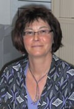 Porträt einer erwachsenen Person mit kurzen dunklen Haaren und Brille. Neutraler Gesichtsausdruck, Blick zur Kamera. Die Person trägt eine gemusterte Bluse und sitzt in einem Innenraum.