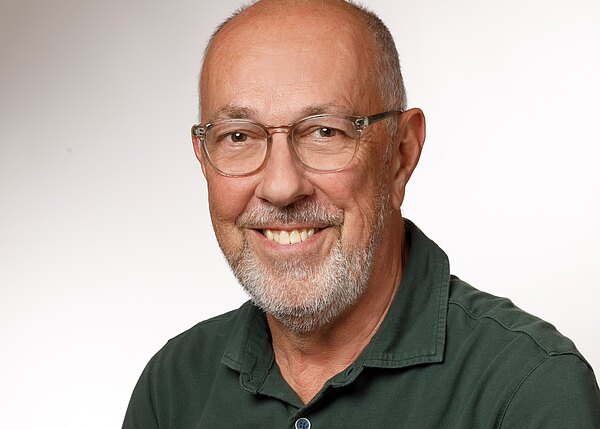 Porträt eines älteren Mannes, der freundlich in die Kamera lächelt. Er trägt Brille, kurzen grauen Bart und ein grünes Polohemd. Heller, neutraler Hintergrund; Fokus auf ruhigen, sympathischen Gesichtsausdruck.