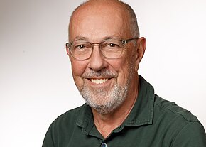 Porträt eines älteren Mannes, der freundlich in die Kamera lächelt. Er trägt Brille, kurzen grauen Bart und ein grünes Polohemd. Heller, neutraler Hintergrund; Fokus auf ruhigen, sympathischen Gesichtsausdruck.