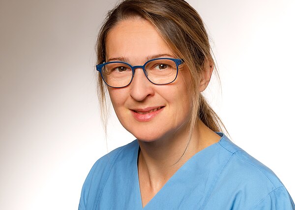 Porträt einer lächelnden Frau mit blauer Brille in blauer medizinischer Kleidung vor neutralem Hintergrund.