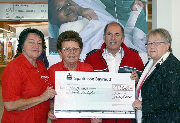 Vier Erwachsene in Rotkreuz-Kleidung präsentieren einen übergroßen Spendenscheck der Sparkasse Bayreuth über 500 Euro. Der Scheck ist für Hilfe in Syrien bestimmt. Sie stehen zusammen und lächeln in die Kamera; im Hintergrund ein Krankenhausplakat.