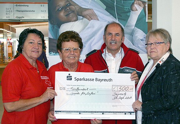 Vier Erwachsene in Rotkreuz-Kleidung präsentieren einen übergroßen Spendenscheck der Sparkasse Bayreuth über 500 Euro. Der Scheck ist für Hilfe in Syrien bestimmt. Sie stehen zusammen und lächeln in die Kamera; im Hintergrund ein Krankenhausplakat.