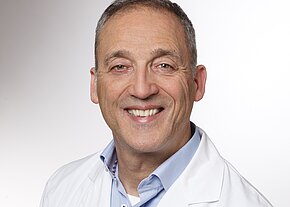 Porträt eines lächelnden Mannes mittleren Alters in weißem Kittel und hellblauem Hemd vor neutralem Hintergrund; vermittelt Freundlichkeit und Professionalität.