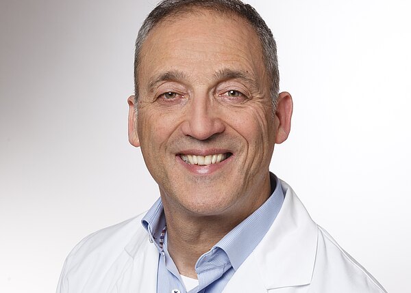 Porträt eines lächelnden Mannes mittleren Alters in weißem Kittel und hellblauem Hemd vor neutralem Hintergrund; vermittelt Freundlichkeit und Professionalität.