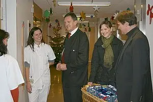 Vier Personen stehen in einem Flur eines Krankenhauses. Eine Krankenschwester in Uniform lächelt, während drei Gäste, darunter zwei Frauen und ein Mann, mit einem Korb voller Geschenke ankommen. Im Hintergrund ist ein Christbaum und weihnachtliche Dekoration zu sehen.