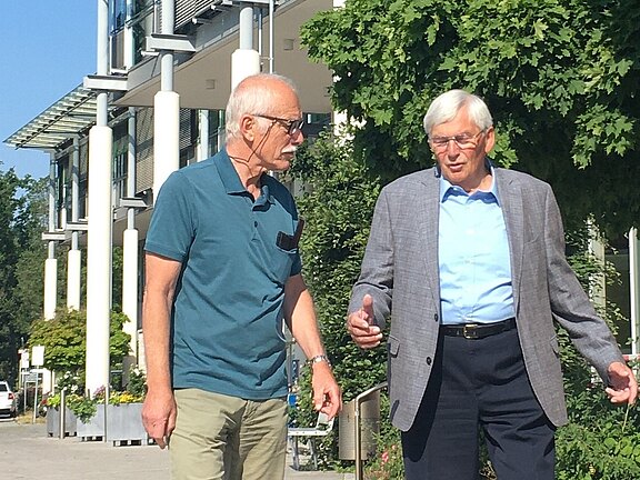 Zwei ältere Männer gehen draußen nebeneinander und unterhalten sich. Der linke trägt ein türkisfarbenes Poloshirt und Sonnenbrille, der rechte ein hellblaues Hemd mit grauem Sakko und gestikuliert beim Sprechen. Im Hintergrund ein modernes Gebäude und Bäume, sonniges Wetter.