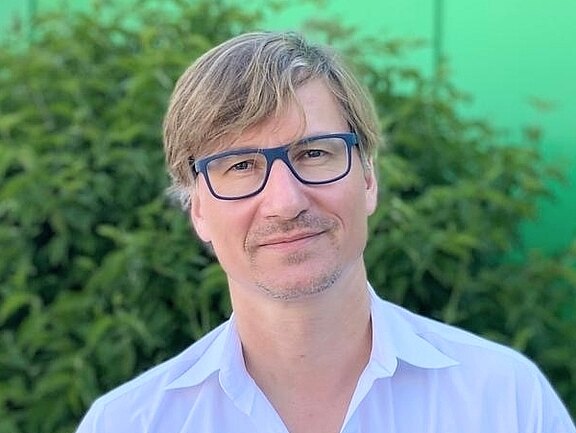 Ein Mann mit blonden Haaren und Brille steht vor einem grünen Hintergrund. Er trägt ein weißes Hemd und hat einen neutralen Ausdruck im Gesicht. Im Hintergrund sind blühende Pflanzen sichtbar.