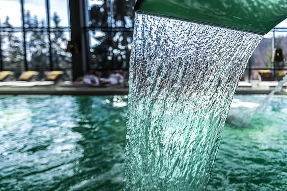 Nahaufnahme eines Wasserfalls aus einer Metallrinne, der in ein türkisfarbenes Hallenbecken stürzt; im Hintergrund unscharf Liegen und große Fenster.