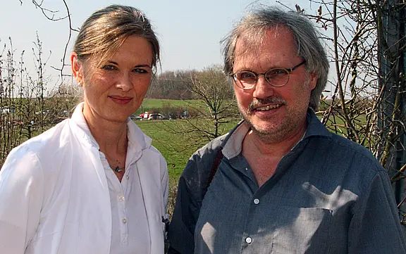 Zwei Erwachsene stehen im Freien nebeneinander. Links eine Frau in weißem Mantel, lächelnd; rechts ein Mann mit Brille und grauem Hemd, ebenfalls lächelnd. Heller Sonnenschein, unscharfe Wiese und Bäume im Hintergrund.