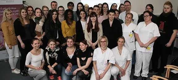 Eine Gruppe von etwa 30 Personen steht in einem Raum. Die Mehrheit trägt alltägliche Kleidung, einige tragen weiße Arbeitskleidung. Im Hintergrund sind Banner und eine Präsentation sichtbar. Die Stimmung wirkt freundlich und gemeinschaftlich.