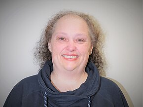 Iris Kammerer Porträt einer lächelnden Person mit schütterem, lockigem Haar. Sie trägt einen dunklen Kapuzenpullover und schaut direkt in die Kamera vor einem neutralen, hellen Hintergrund.